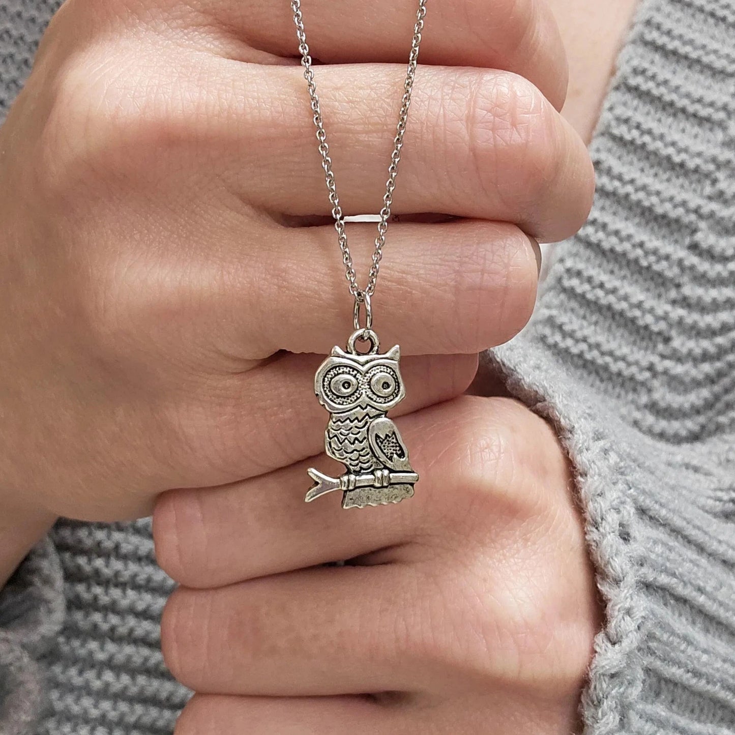 Silver owl pendant necklace held by a hand against a gray sweater background