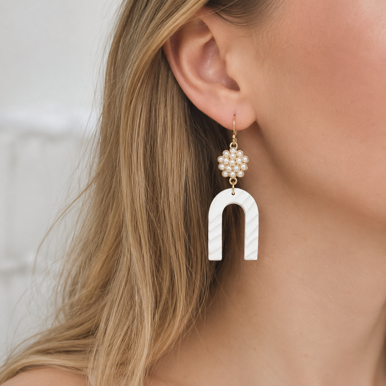 A close-up side profile photograph of a blonde model wearing a statement dangle earring featuring a gold flower motif cluster of tiny pearls and a large, textured white U-shaped arch.