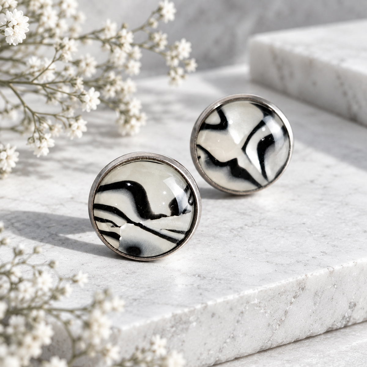 Black and white zebra print round stud earrings with resin domes and stainless steel posts, displayed on white stone with delicate flowers.