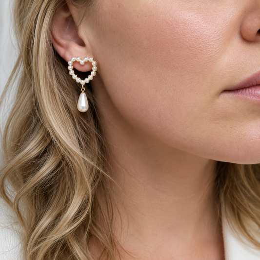 A model with blonde hair wearing a gold heart-shaped pearl drop earring featuring small white faux pearls and a smooth pearl teardrop dangle accessory.