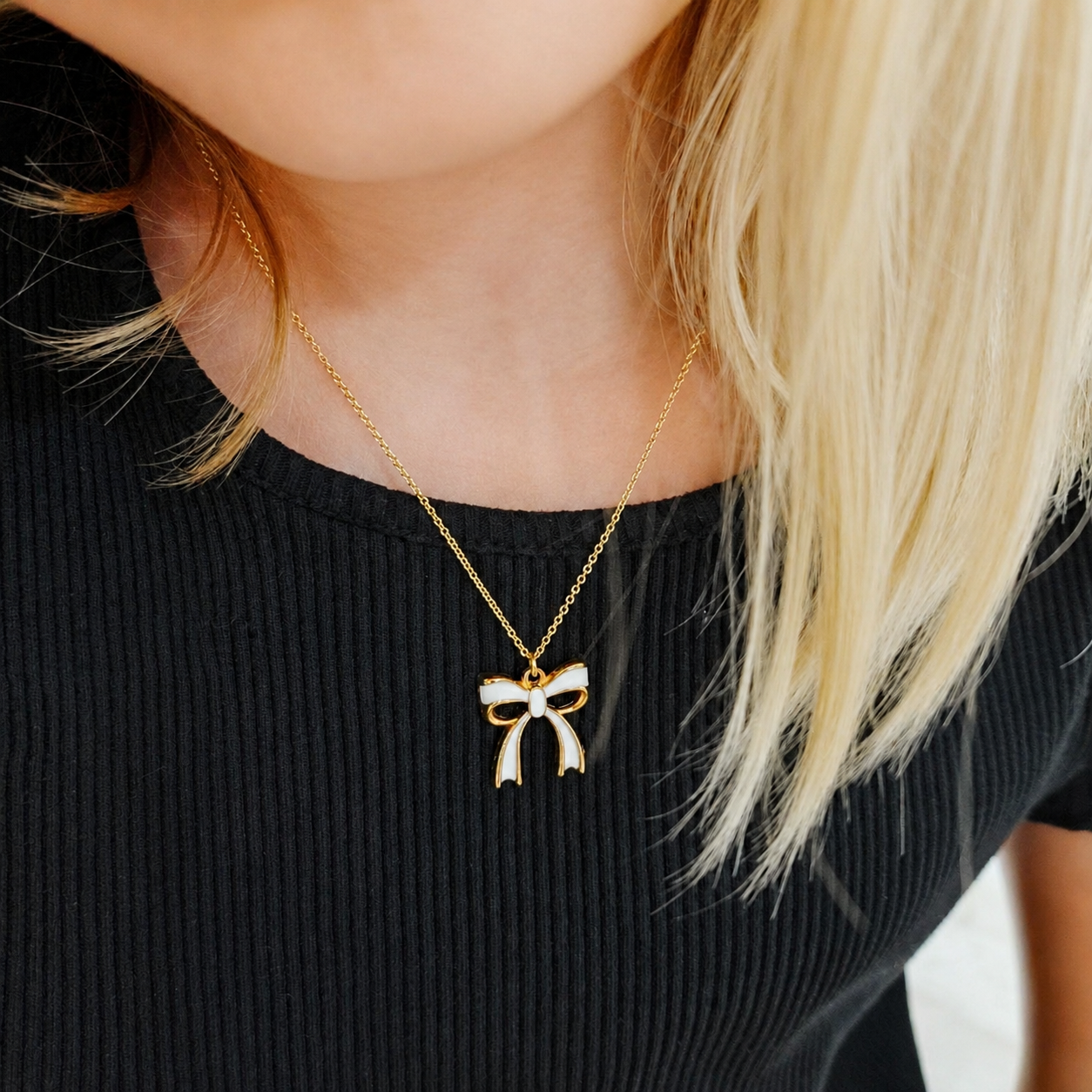 A blonde model wearing a dainty gold chain necklace featuring a white enamel bow pendant with a gold border, styled over a ribbed black top.