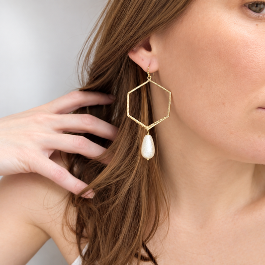 Close-up of a woman modeling a long, gold hexagon-shaped earring with a single teardrop pearl pendant. Her hand is gently tucked into her hair, showcasing the earring’s length and elegant design against a soft, neutral background.
