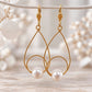 Gold teardrop pearl earrings hanging against a soft neutral floral background with textured gold wire and white pearl accents.