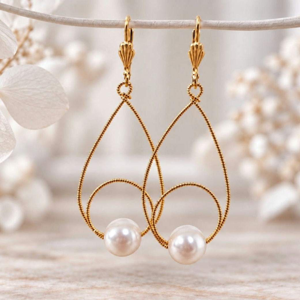 Gold teardrop pearl earrings hanging against a soft neutral floral background with textured gold wire and white pearl accents.