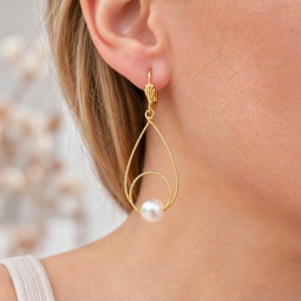 Close-up of a blonde woman wearing a gold teardrop pearl earring with a decorative leverback hook.