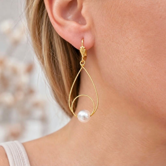Close-up of a blonde woman wearing a gold teardrop pearl earring with a decorative leverback hook.