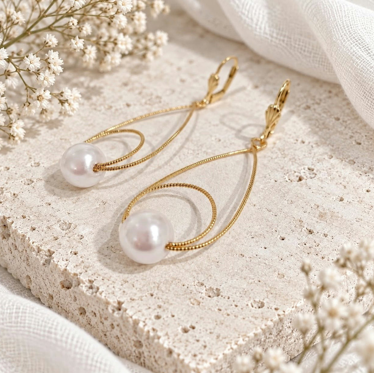 Gold teardrop pearl earrings displayed on pale ivory textured stone with white flowers and soft neutral fabric.