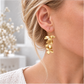 Close-up of a blonde woman wearing a long gold orchid earring with cascading brushed metal flowers, styled with swept-back hair and a white textured top.