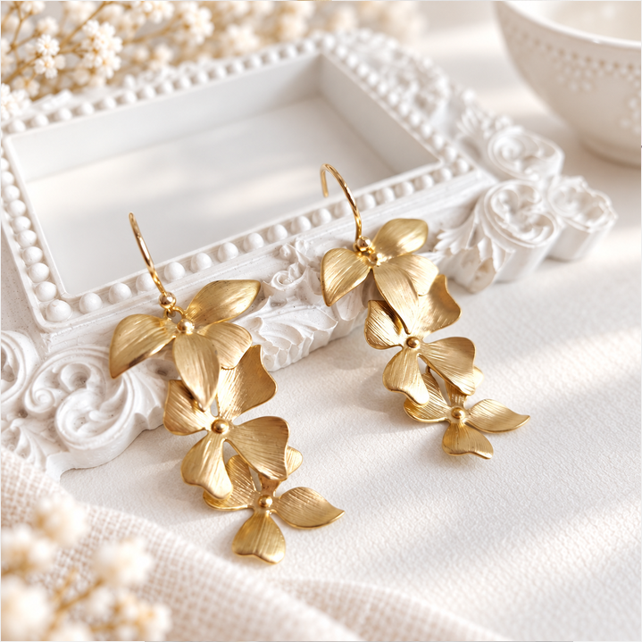 Pair of cascading gold floral earrings arranged on an ornate white frame with decorative scrollwork, surrounded by soft white flowers and neutral fabric.