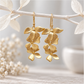 Pair of long cascading gold orchid earrings hanging from a delicate white branch, featuring layered matte gold floral petals with a brushed metal texture and gold-plated ear wires against a soft neutral background.