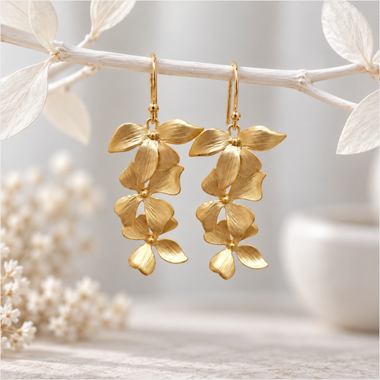 Pair of long cascading gold orchid earrings hanging from a delicate white branch, featuring layered matte gold floral petals with a brushed metal texture and gold-plated ear wires against a soft neutral background.