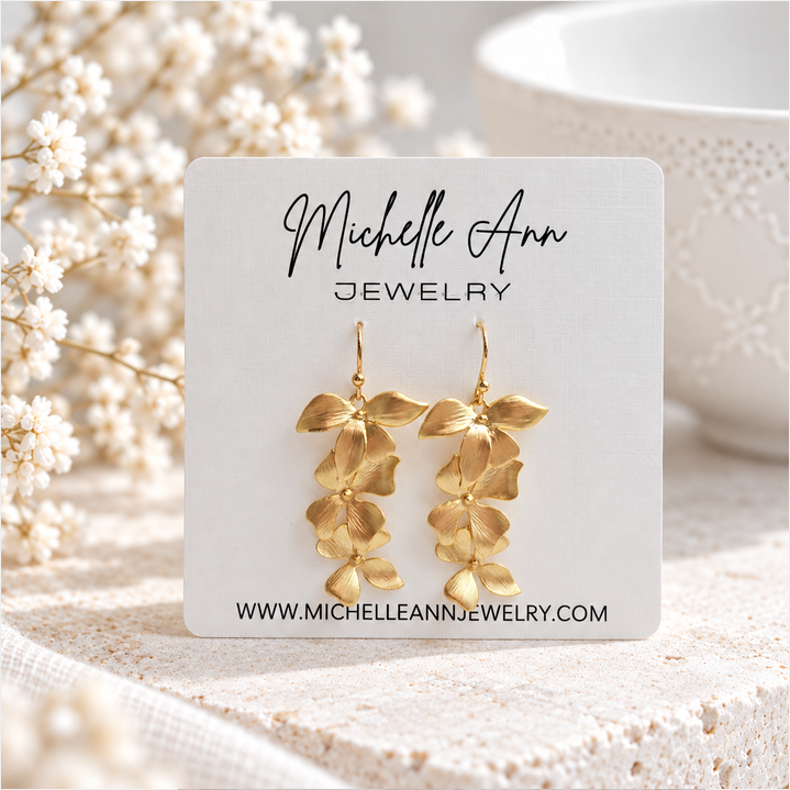 Gold orchid dangle earrings displayed on a Michelle Ann Jewelry card, surrounded by delicate white flowers, with a soft neutral background and pale stone surface.