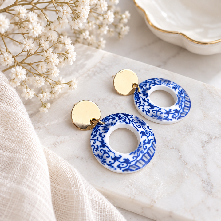 Blue and white chinoiserie-inspired acrylic resin earrings with gold-plated studs styled on white marble with delicate flowers and ivory fabric.