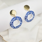 Blue and white floral acrylic resin dangle earrings with round gold-plated studs, displayed on a white jewellery stand.