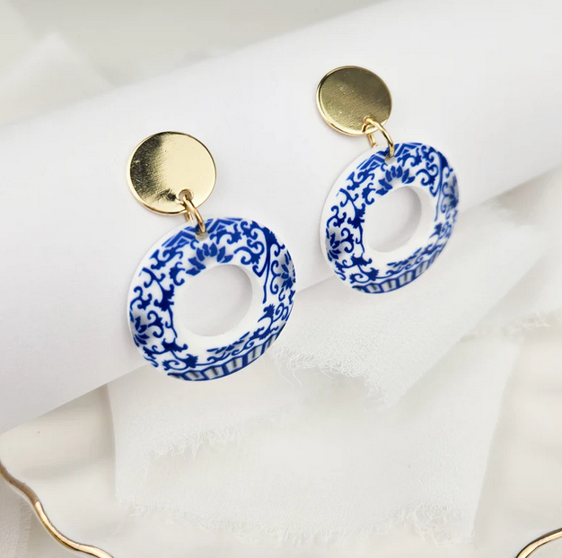 Blue and white floral acrylic resin dangle earrings with round gold-plated studs, displayed on a white jewellery stand.