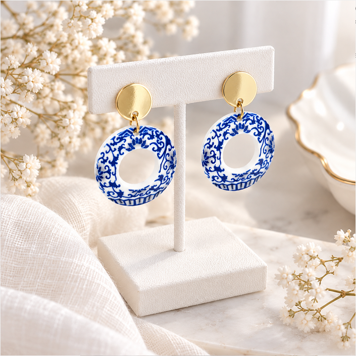 A pair of dangling hoop earrings displayed on a white jewelry stand. The hoops are blue and white patterned with an intricate floral design, suspended from round gold-tone posts, set against a soft, styled background of white flowers and neutral fabric.