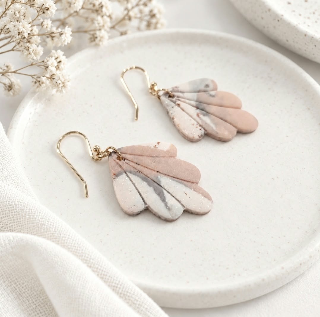 Blush pink, ivory and grey marbled polymer clay earrings with delicate gold accents arranged on a cream ceramic dish.