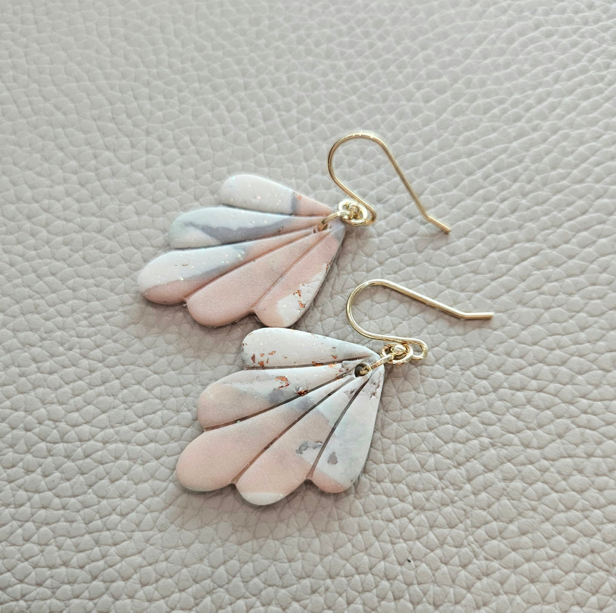 Close-up of handmade polymer clay fan earrings featuring soft pink, white and grey marbling, subtle metallic flecks and gold-tone hooks.