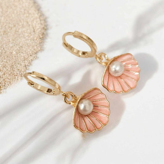 Close-up of pink seashell earrings with pearl accents and gold hardware, styled on a white surface with soft beach sand.