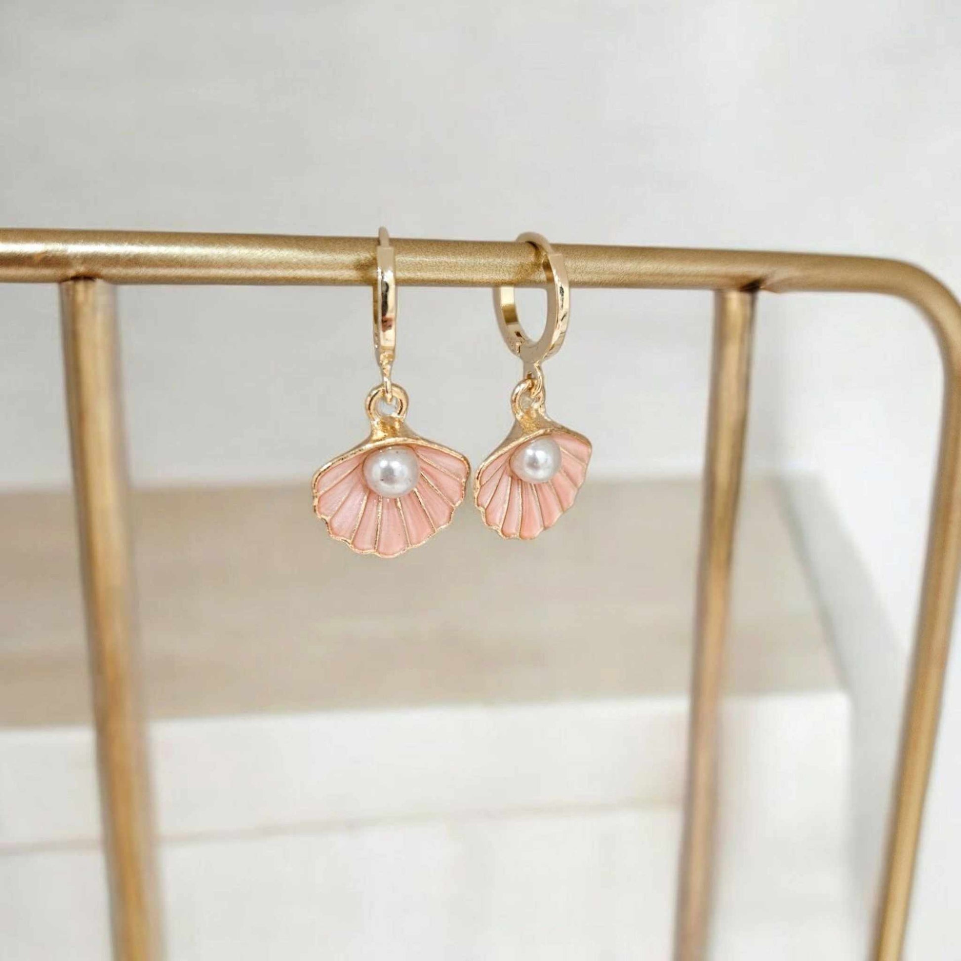 Elegant pink shell and pearl dangle earrings hanging on a gold rack, showcasing their delicate ocean-inspired design.