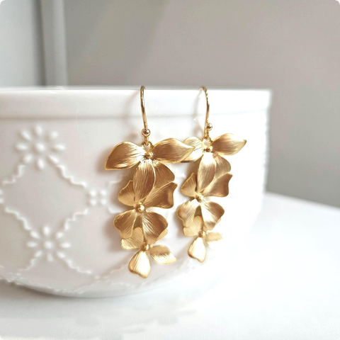 Cascading matte gold orchid earrings hanging from a white embossed ceramic bowl, featuring layered floral petals with brushed metal texture and gold-plated ear wires.