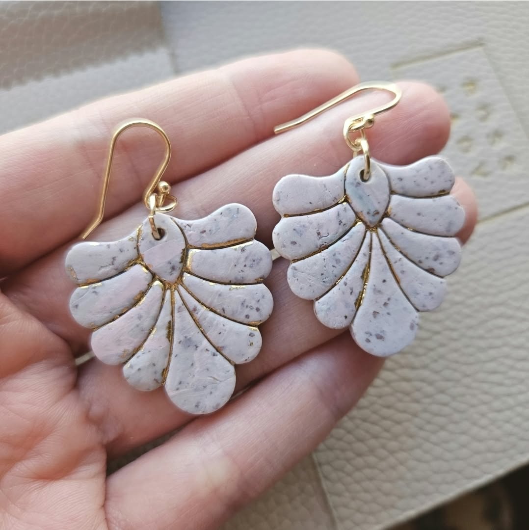 Hand holding blush pink and ivory speckled fan-shaped polymer clay earrings with gold hooks and gold detailing.