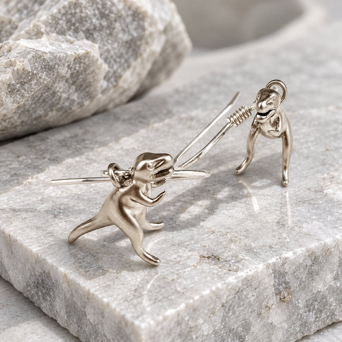 T-Rex dangle earrings with sterling silver ear wires displayed on grey stone.
