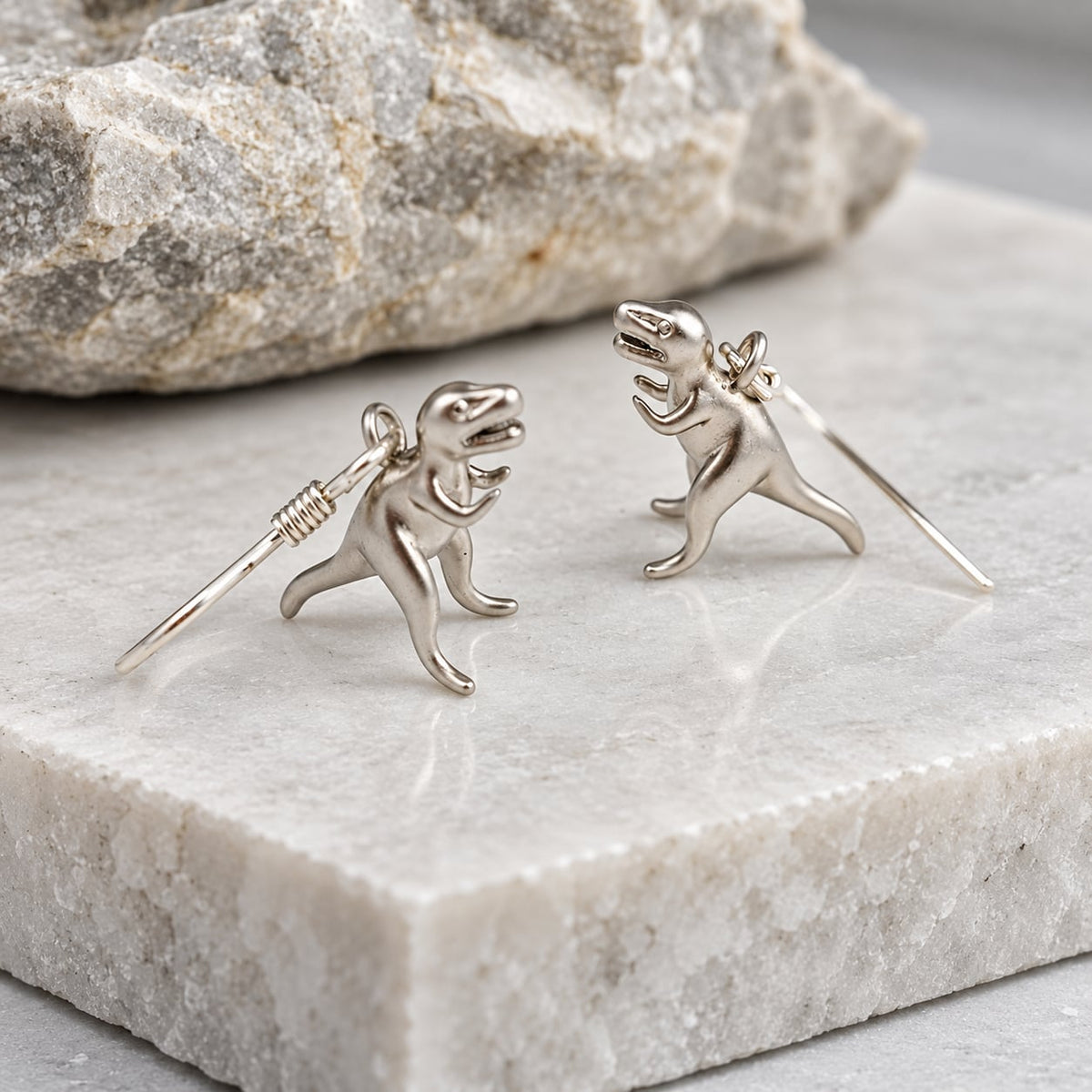 Silver T-Rex dinosaur earrings standing on a textured grey stone display.