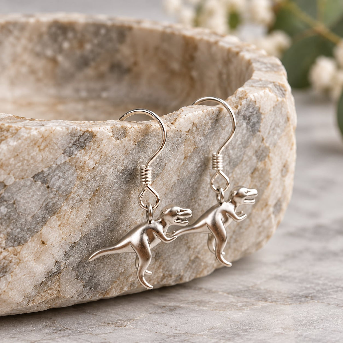 T-Rex dangle earrings with sterling silver ear wires hanging from a natural stone edge.