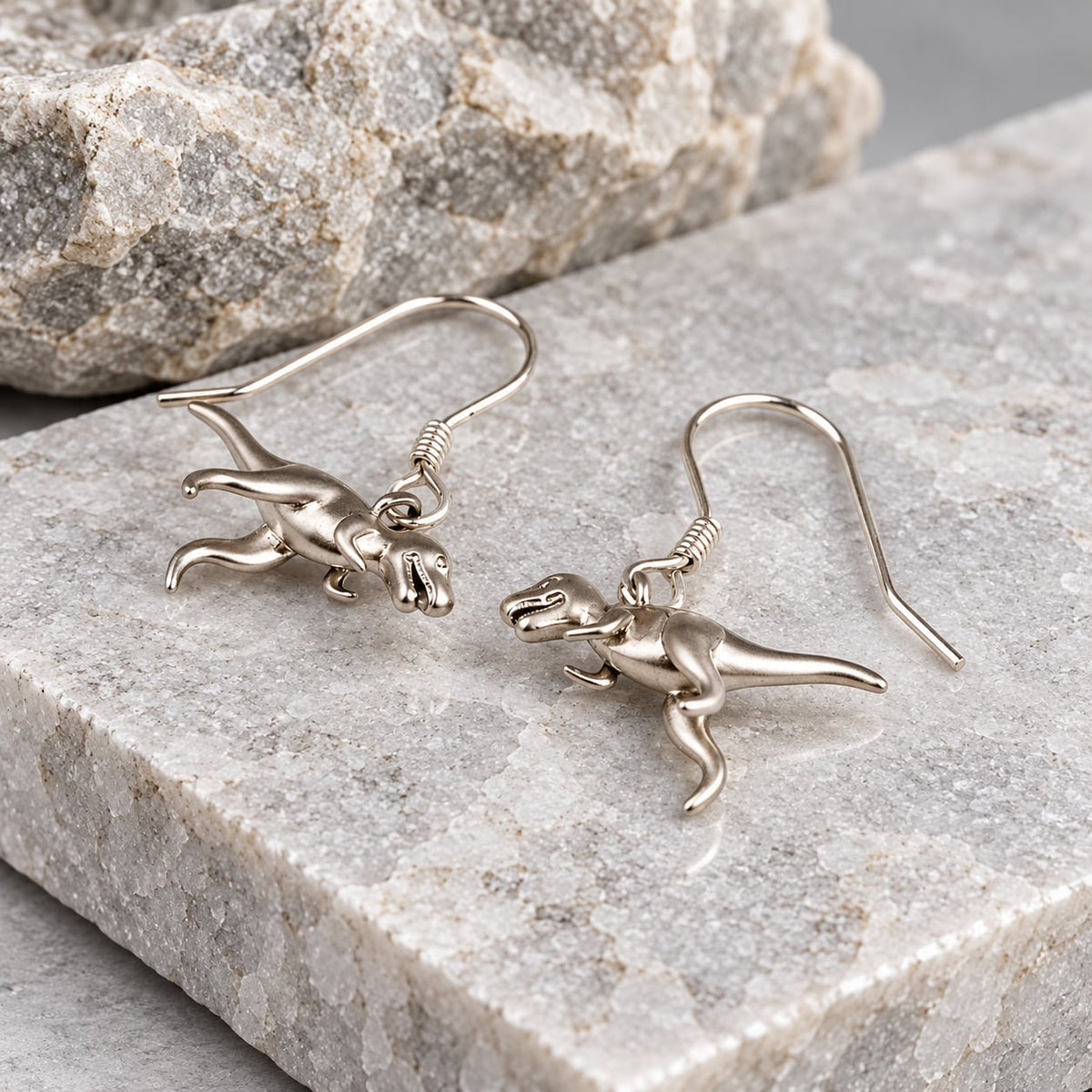 Silver T-Rex dinosaur earrings shown from the side on a grey stone surface. 