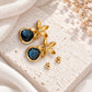 Gold floral drop earrings with dark smoky blue faceted teardrop stones displayed on pale ivory textured stone with white flowers.