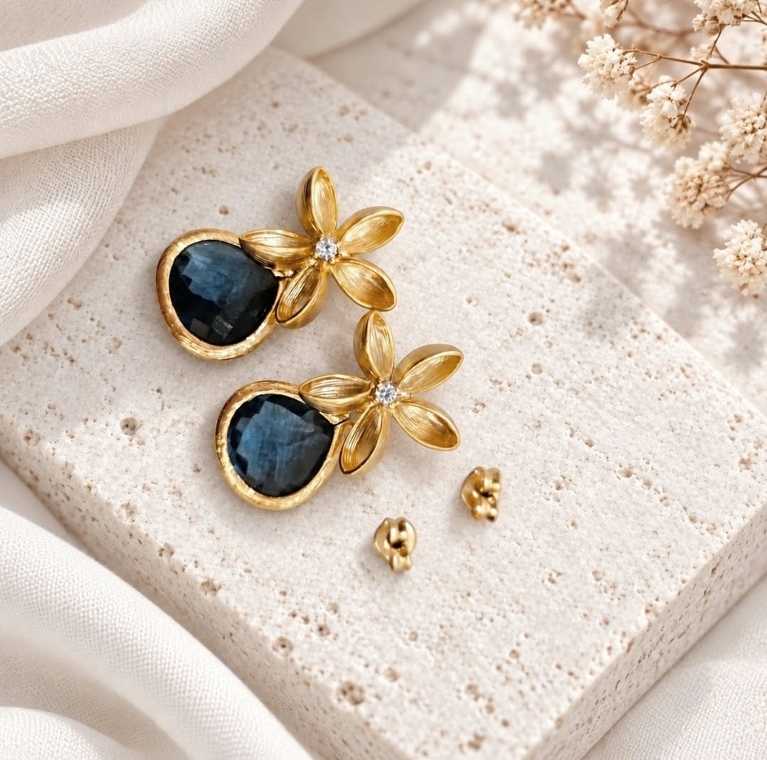 Gold floral drop earrings with dark smoky blue faceted teardrop stones displayed on pale ivory textured stone with white flowers.