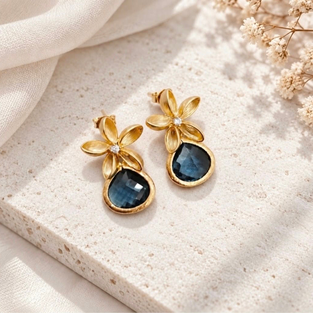 Gold flower earrings with deep blue teardrop stones styled on a light cream stone surface with soft ivory fabric and dried flowers.