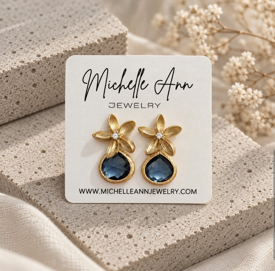 Gold floral blue stone earrings displayed on a Michelle Ann Jewelry card against neutral textured stone and soft white floral accents.