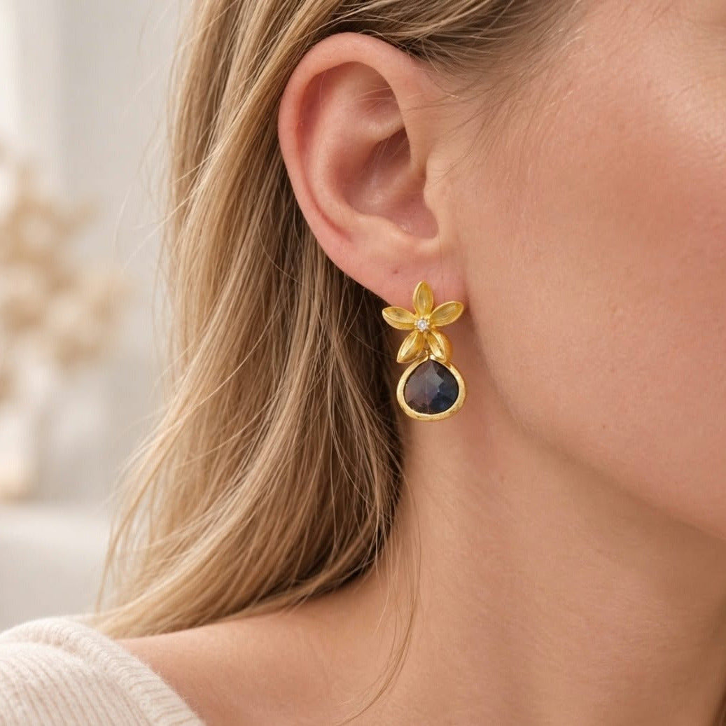 Close-up of a blonde woman wearing a gold flower drop earring with a dark blue faceted teardrop stone.
