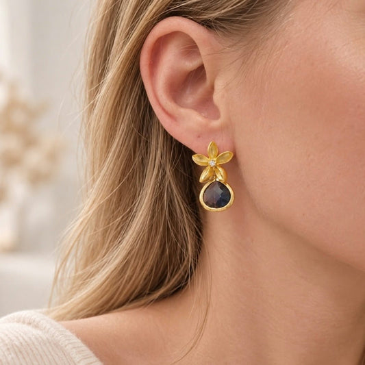 Close-up of a blonde woman wearing a gold flower drop earring with a dark blue faceted teardrop stone.