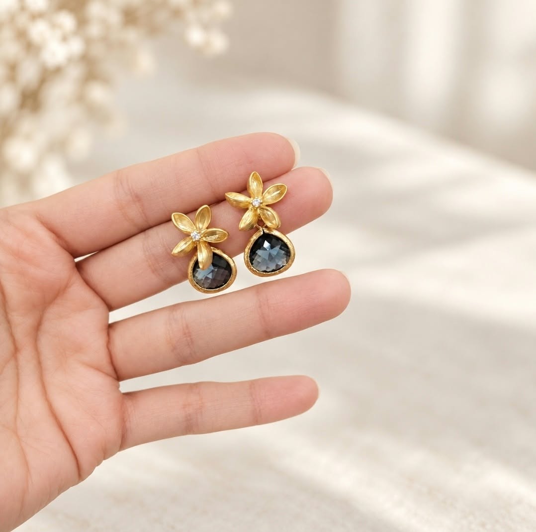 Hand holding a pair of gold floral earrings with smoky blue faceted teardrop stones against a soft neutral cream background.
