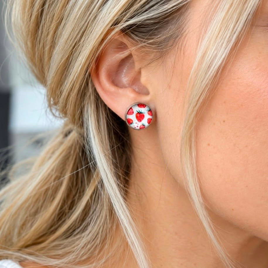A close-up of a woman's ear wearing a round stud earring with a red and white heart pattern on a white background.