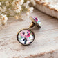 Close-up of round antique bronze stud earrings featuring pink wildflowers under glass domes, showing the earring posts and raised glass setting.