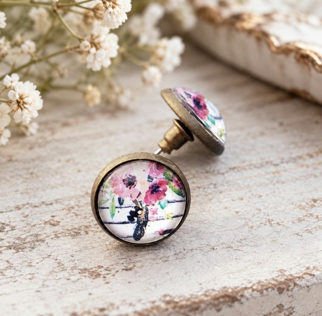 Close-up of round antique bronze stud earrings featuring pink wildflowers under glass domes, showing the earring posts and raised glass setting.
