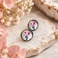 Pair of antique bronze floral stud earrings with pink, green and black wildflower artwork, styled with soft blush and white dried flowers.