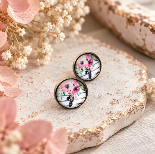 Pair of antique bronze floral stud earrings with pink, green and black wildflower artwork, styled with soft blush and white dried flowers.