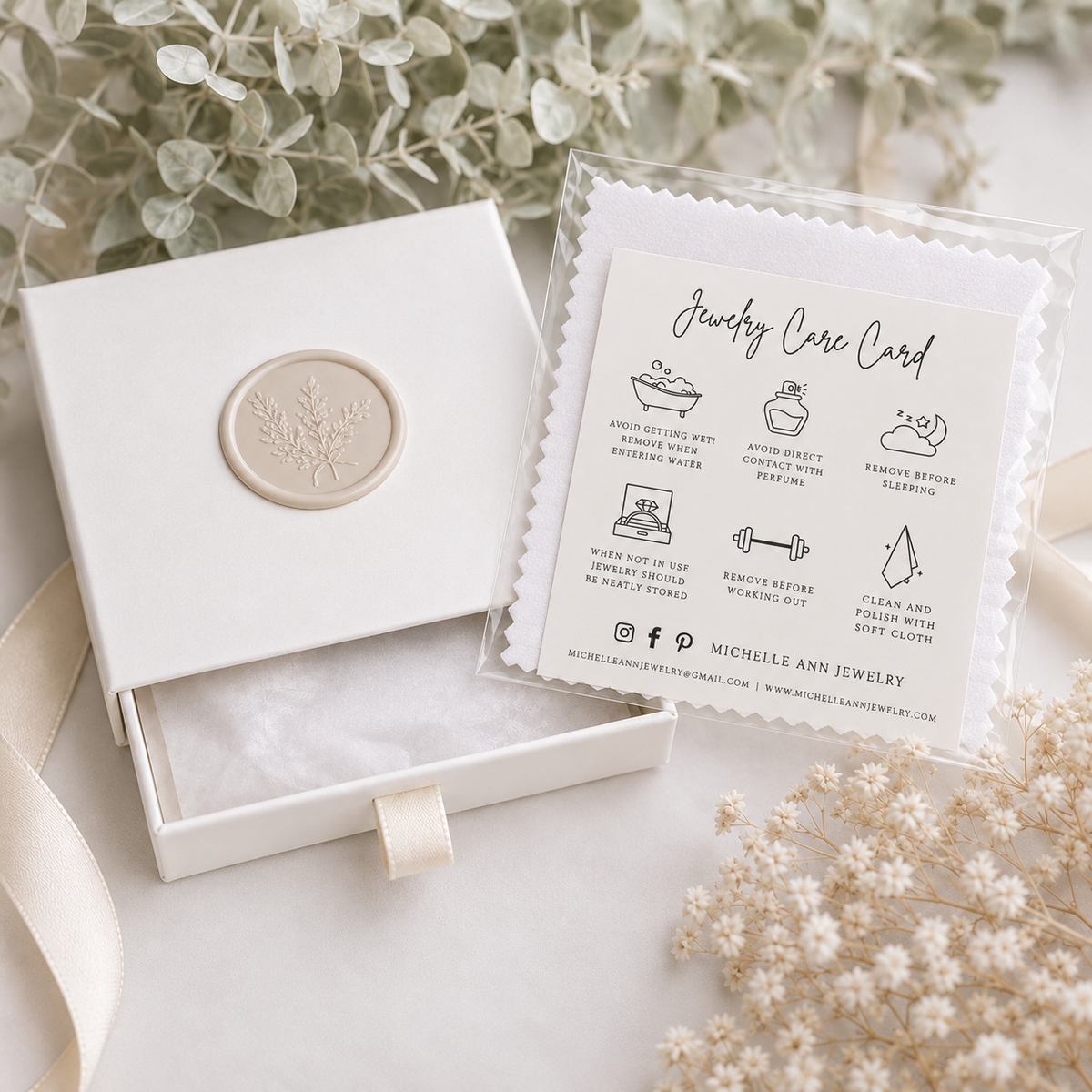 White Michelle Ann Jewelry gift box with cream botanical seal, polishing cloth and jewelry care card, styled with soft neutral flowers and ribbon.