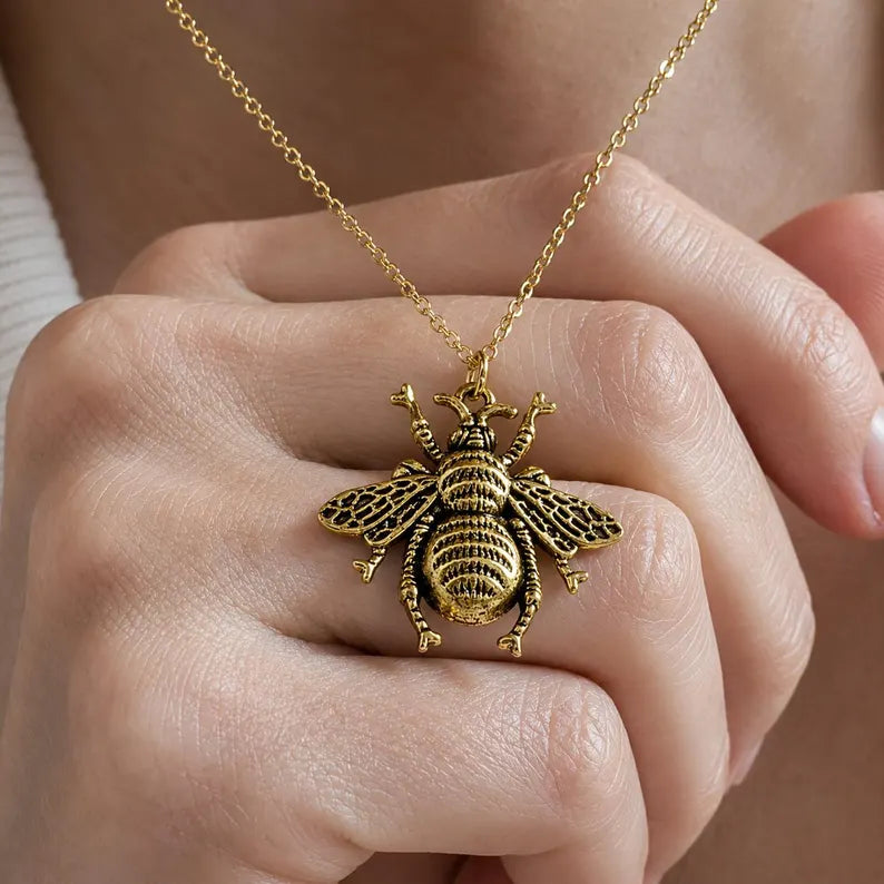 Close-up of a woman holding a detailed gold bee necklace at her neckline, styled with a white sweater in natural light.