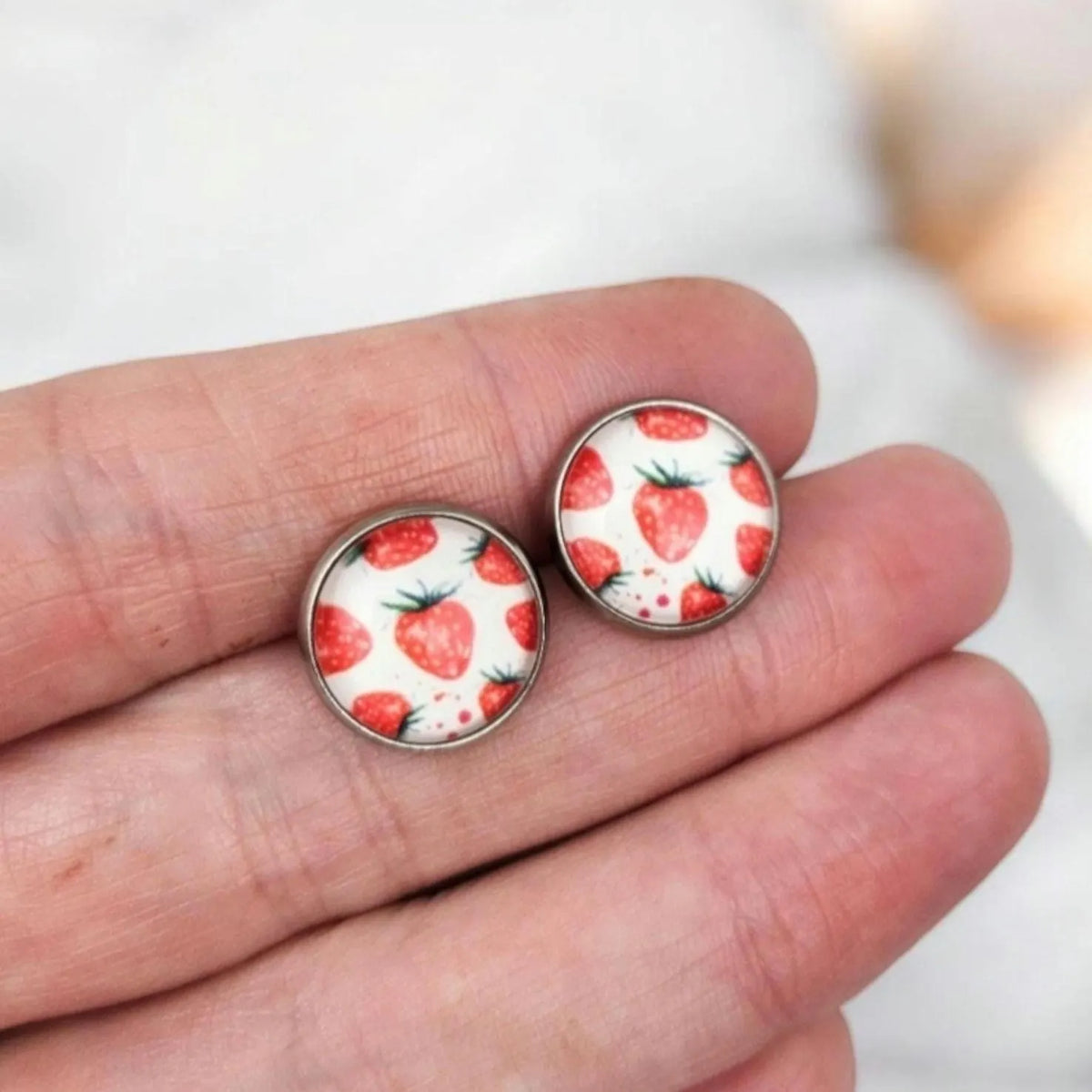 Silver strawberry stud earrings featuring hypoallergenic stainless steel posts and a cute strawberry glass dome design. Lightweight for comfortable all-day wear. Hand size comparison.