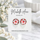 A pair of stud earrings with a red strawberry pattern on a white background, mounted on a white display card for Michelle Ann Jewelry, styled on a marble surface with white flowers.