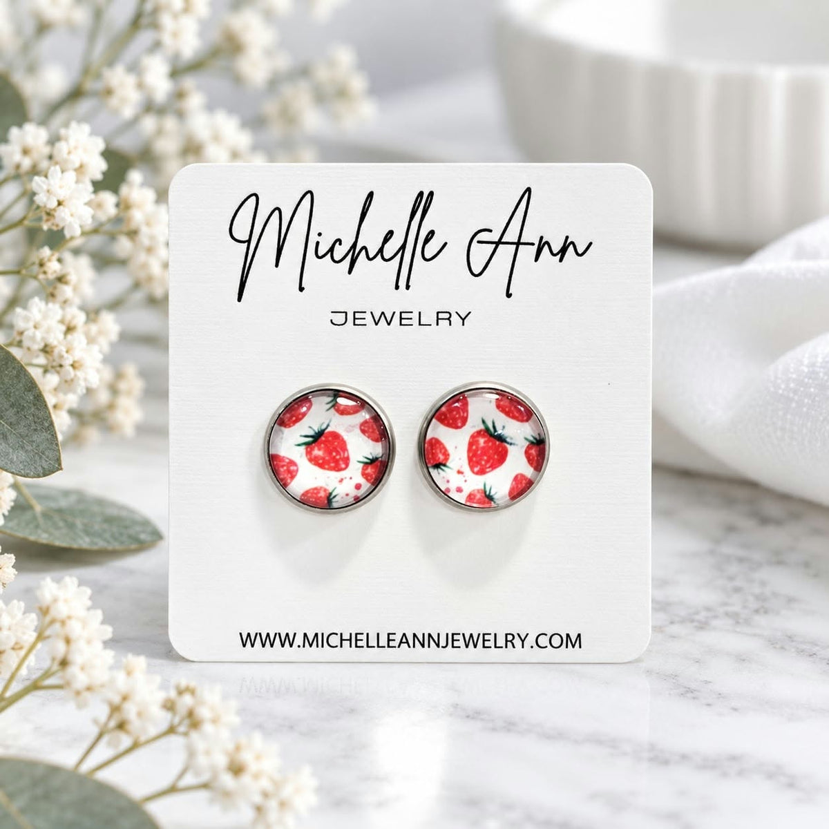 A pair of stud earrings with a red strawberry pattern on a white background, mounted on a white display card for Michelle Ann Jewelry, styled on a marble surface with white flowers.