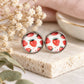 A pair of round stud earrings with a white background and a red strawberry pattern, set in a silver-tone metal frame and displayed on a textured stone block with dried flowers.