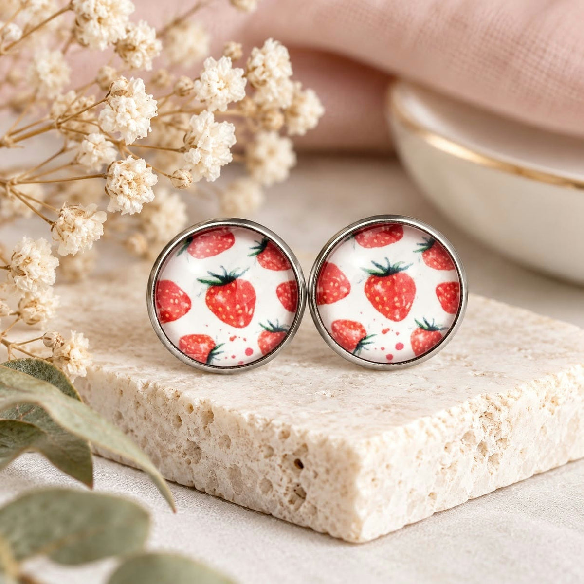 A pair of round stud earrings with a white background and a red strawberry pattern, set in a silver-tone metal frame and displayed on a textured stone block with dried flowers.